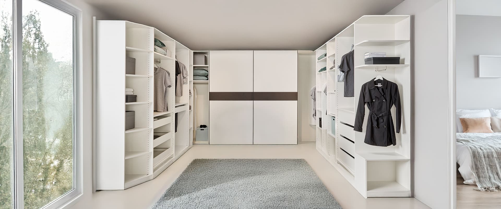 Ankleidezimmer über zwei Ecken in weiß. Der Schwebetürenschrank hat eine abnehmbare Blende in Schiefergrau Dekor und die Kleiderschränke haben keine Türen.