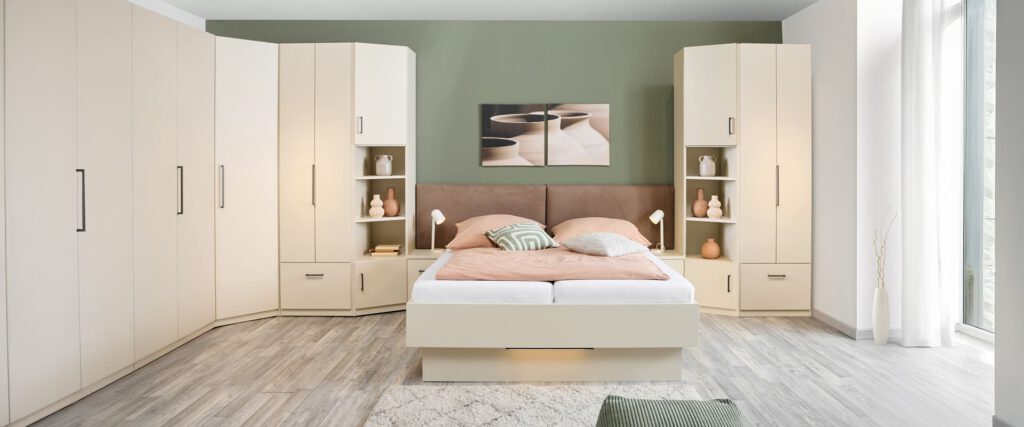 Schlafzimmerschränke aus der Serie MULTIFLEX BASIC. Die Kombination geht über Eck. Mittendrin ein Studio Bett mit seitlichen Nachtkonsolen und zwei stoffbezogenen Kopfteilen. Komplette Wand in der Farbe sand
