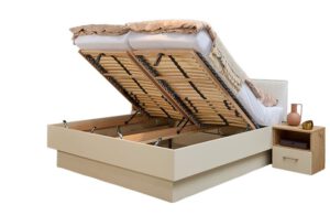 Studio Bett mit zwei geöffneten Doppelbett Studio mit stoffbezogenem Kopfteil und einer Nachtkonsole DeLuxe. Man blickt etwas in den Stauraum. Rechts daneben steht eine Nachkonsolte mit einem Schubkasten und einem offenen Fach