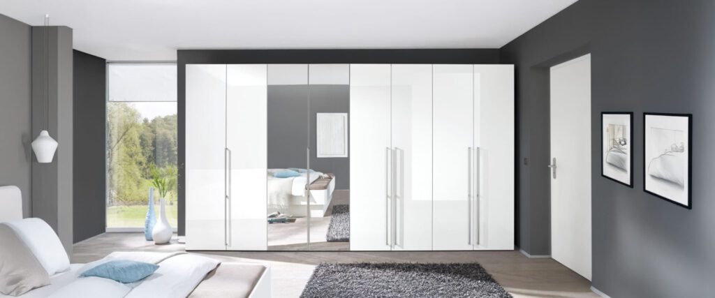 Kleiderschrankzeile in weiß mit langen Stangengriffen. Die rechte Falttür von links ist statt Holz vollflächig mit einem Spiegel ausgestattet